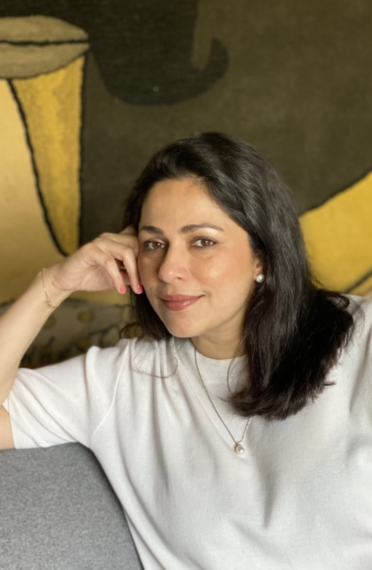 Divya C. Berry, psychologist and Gestalt therapist, seated in a white t-shirt against a patterned wall hanging.